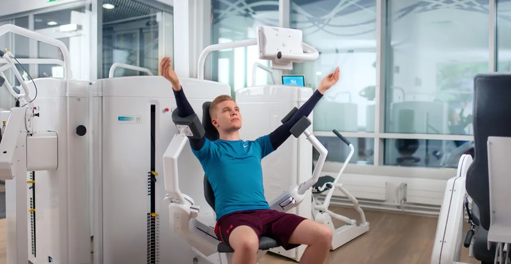 Patientin trainiert an einem Schultertherapiegerät mit Bildschirmanzeige